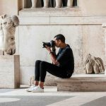 a photographer taking pictures sitting on a stair with a marble statue nearby