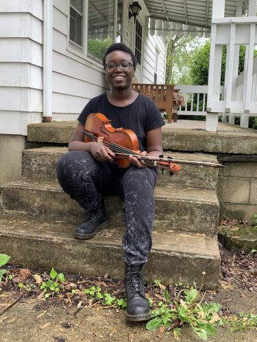 a young women with a violin sits on stone stairs in front on the porch of a white house.