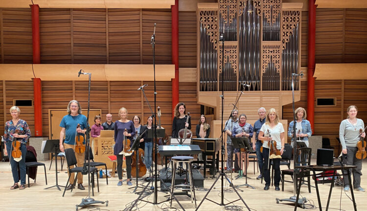 members of an orchestra stand with instruments among microphones in a room with a large organ in the background.