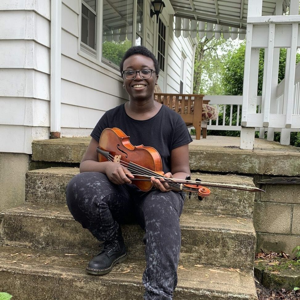 a young women with a violin sits on stone stairs in front on the porch of a white house.