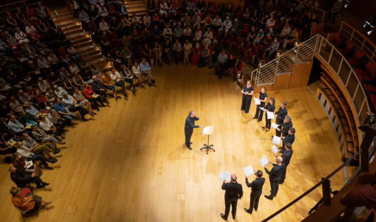 A man stands on a stage with a music stand in front of him. directly in front of him are singers with music in a semi-circle. Behind is a large audience in a semi-circle.