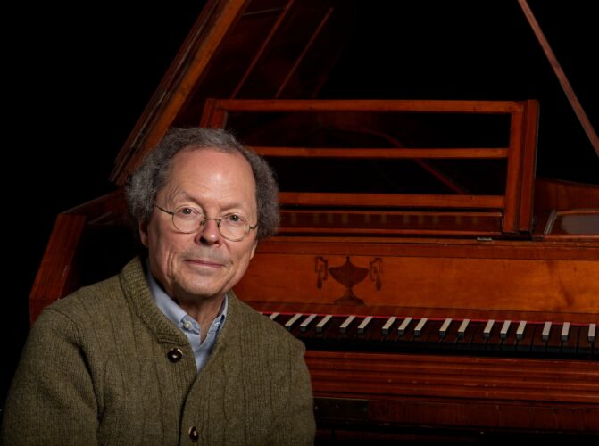 a man with glasses in a green cartigan sits in front of a harpsichord and a black background.